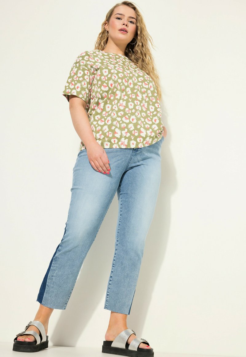Zelená květinová potisková blůza s krátkým rukávem, spárovaná se světle modrými džínami po kotníky a stříbrnými sandály s tlustými černými podrážkami.