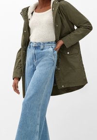 Olivově zelená parka s chlupatou podšívkou, kombinovaná s širokými světle modrými džíny. Parka má zip a kapsy.