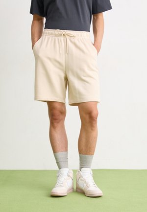 Person iført beige shorts med snøre, grå sokker, hvide høje sneakers og en mørk t-shirt, stående på en grøn gulv mod en hvid væg.