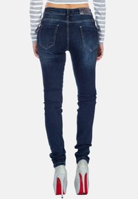 Jeans skinny in lavaggio scuro con tasche posteriori, chiusura con bottone e una leggera scoloritura. Abbinati a tacchi alti argento con accenti rossi.