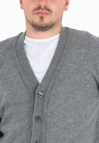 Cardigan grigio a maglia con scollo a V, dotato di cinque bottoni e una finitura testurizzata. Indossato sopra una maglietta bianca semplice.