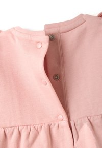 Primer plano de una prenda de bebé rosa con broches a presión a lo largo del cuello en la parte posterior y tela fruncida en la cintura.