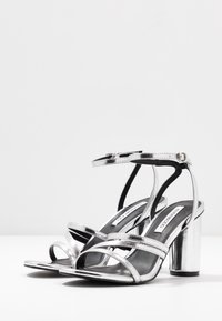 Srebrne sandały na wysokim obcasie z kilkoma skrzyżowanymi paskami, masywnym metalicznym obcasem blokowym i błyszczącym wykończeniem z materiału syntetycznego.