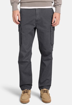 Mand iført mørkegrå cargobukser med sidelommer og beige snørestøvler, stående mod en ensfarvet baggrund.