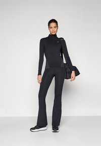 Zwarte sportieve lange mouw top met hoge kraag en rits, gecombineerd met bijpassende zwarte flared leggings. Model draagt een zwarte sporttas.