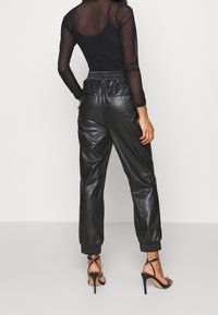 Jogging en cuir noir avec une taille élastique, chevilles resserrées et deux poches arrière, associés à un haut transparent à manches longues noir et des sandales à talons hauts.