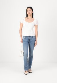 Blouse blanche à manches bouffantes avec des détails texturés, associée à un jean ajusté bleu clair et des ballerines beiges. Modèle tenant un petit sac à main blanc.