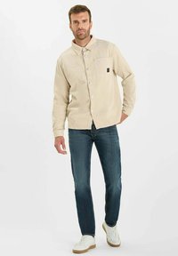 Beige corduroy overhemdjack met een kraag, twee voorkruppen en witte knopen, gecombineerd met donkerblauwe jeans en witte sneakers.