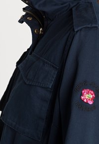 Granatowa parka z kapturem, z kieszenią na piersi, haftowanym wzorem kwiatowym na rękawie oraz złotymi akcentami.