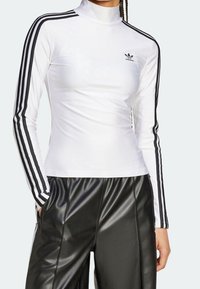 Långärmad, vit idrottströja gjord av stretchigt material med hög krage, med svarta ränder längs ärmarna och en liten logotyp på bröstet.