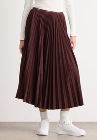 Jupe plissée bordeaux en tissu texturé, avec une taille élastique portant un logo, associée à des baskets blanches et des chaussettes.