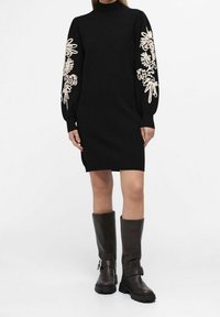 Femme portant une robe noire à manches longues avec une broderie florale beige sur les manches et des bottes marron foncé à boucle jusqu'au genou.