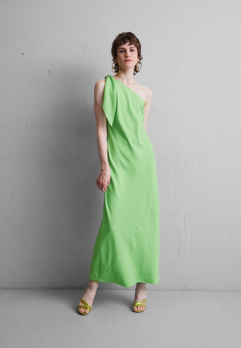 Πράσινο φόρεμα ενός ώμου με ρέουσα σχεδίαση και ομαλή υφή. Σημαντική διακόσμηση με φιόγκο στον ώμο. Κίτρινες σανδάλι με λουράκια.