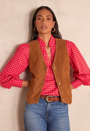 Gilet en daim marron avec deux poches avant, porté sur un chemisier rouge à motifs avec des manches bouffantes. Le modèle porte une ceinture marron et un jean en denim.