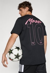 Sort t-shirt med pink tekst "Messi" og nummer "10" på ryggen. Modellen holder en sort-hvid fodbold med stjernemønstre.