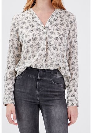 Kvinde iført en hvid bluse med lange ærmer med sorte bladmønstre, stukket ned i mørkegrå jeans med høj talje.