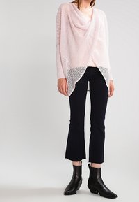 Hellrosa gestrickter Poncho mit einem drapierten Ausschnitt und asymmetrischem Saum, kombiniert mit schwarzen, ausgestellten Jeans und schwarzen Stiefeletten.