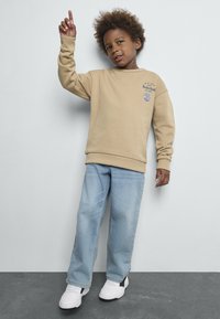 Sweat-shirt beige avec logo, manches longues, col rond ; associé à un jean bleu clair et des baskets blanches avec des accents noirs ; pose debout.