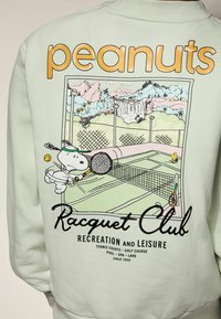 Hellgrüner Sweatshirt mit einem grafischen Druck von Snoopy, der Tennis spielt, sowie dem Text "peanuts" und "Racquet Club" und Landschaftsdetails.