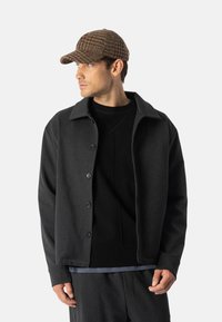 Ciemnoszara kurtka zapinana na guziki z rozłożystym kołnierzem i teksturowanym wełnianym materiałem. Czarny sweter z okrągłym dekoltem pod spodem. Brązowa czapka w deseniu houndstooth.