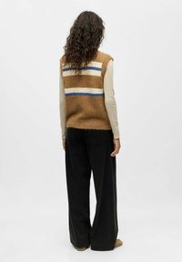 Brązowy sweter w serek z niebieskimi i białymi paskami, noszony na długim rękawie kremowej bluzki, w zestawieniu z czarnymi spodniami wide-leg. Widoczna teksturowana tkanina.