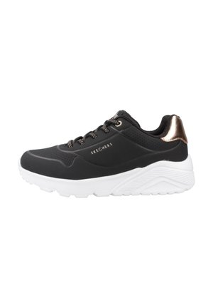 Baskets de sport noires avec une texture lisse, un design perforé, une semelle blanche et un accent en bronze métallisé au niveau du talon. Elles disposent de lacets contrastés.