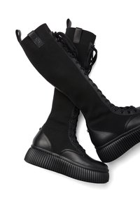 Czarne, wysokie trampki posiadają żeberkową dzianinę oraz gładkie skórzane akcenty, z grubą, teksturowaną podeszwą i sznurówkami sięgającymi do łydki.