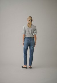 Kobieta z blond kucykiem, ubrana w szary, paskowany top z krótkim rękawem, niebieskie dżinsy i czarne mulesy na niskim obcasie, stojąca na neutralnym tle.