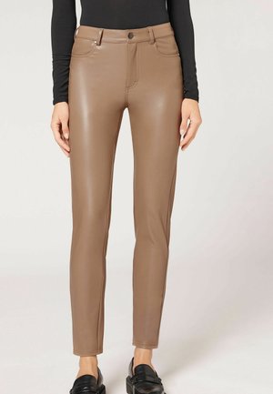 Brązowe wąskie spodnie z ekoskóry z przednimi kieszeniami, noszone z czarnymi mokasynami i czarną bluzką z długim rękawem, pokazane od talii do stóp.