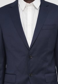 Sacou bleumarin cu două nasturi negri, cu rever în formă de smochin și un buzunar pe piept, purtat peste o cămașă albă cu nasturi, cu guler clasic.