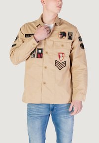 Beige bomuldsjakke med flere lapper, herunder militære insignier og farverige designs. Har knaplukning foran og to brystlommer.