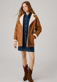 Cappotto in suede marrone con colletto in shearling bianco, dotato di tasche laterali e vestibilità rilassata. Indossato sopra un vestito di denim e stivali da cowboy marroni.