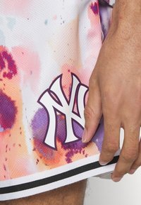 Los pantalones cortos están hechos de una tela de malla ligera y presentan un colorido patrón de tie-dye en tonos de rosa y morado. Un logo de Nueva York en blanco está contorneado en morado.