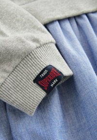 Szürke bordázott gallér, sötétkék címkével, rajta piros "Superdry" felirattal, kék és fehér kockás anyaggal alatta.