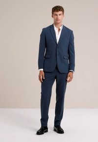 Man står, klädd i en marinblå kostym med en vit skjorta och svarta dressskor framför en enfärgad beige bakgrund.