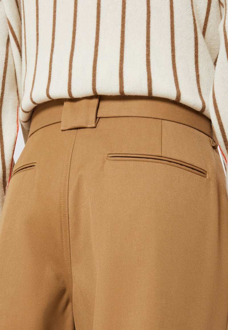 Pantalones marrones a medida con una textura suave, que cuentan con dos bolsillos traseros y una cinturilla sutil. Suéter a rayas blanco y marrón visible en la parte superior.