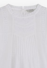 Blusa blanca con intrincados detalles de encaje, frente plisado y cierre en la espalda tipo ojal. La tela tiene una textura suave y ligera.
