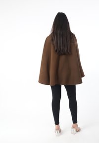 Capa marrón texturizada con una silueta amplia, usada sobre leggings negros. Sandalias transparentes con tacón grueso completan el conjunto.