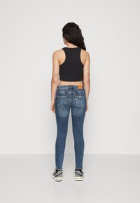 Jeans ajustados en denim azul deslavado con un ajuste de cintura media, combinados con una camiseta de tirantes negra corta. Incluye bolsillos traseros y un parche de logo marrón.