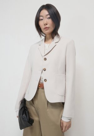 Woman wearing a light beige blazer, white top, khaki pants, holding a black clutch against a plain white background.