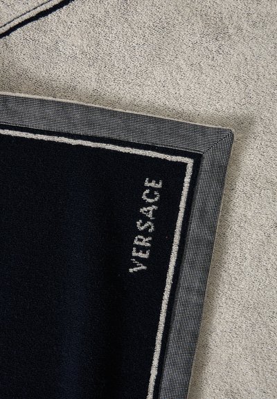 Γωνία μιας μαύρης πετσέτας Versace με λευκό λογότυπο και περίγραμμα, τοποθετημένη σε υφή μπεζ επιφάνειας.