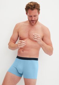 Lichtblauwe katoenen boxershorts met een zwarte elastische tailleband, aansluitend ontwerp en gladde textuur, model getoond met een blote bovenlichaam.