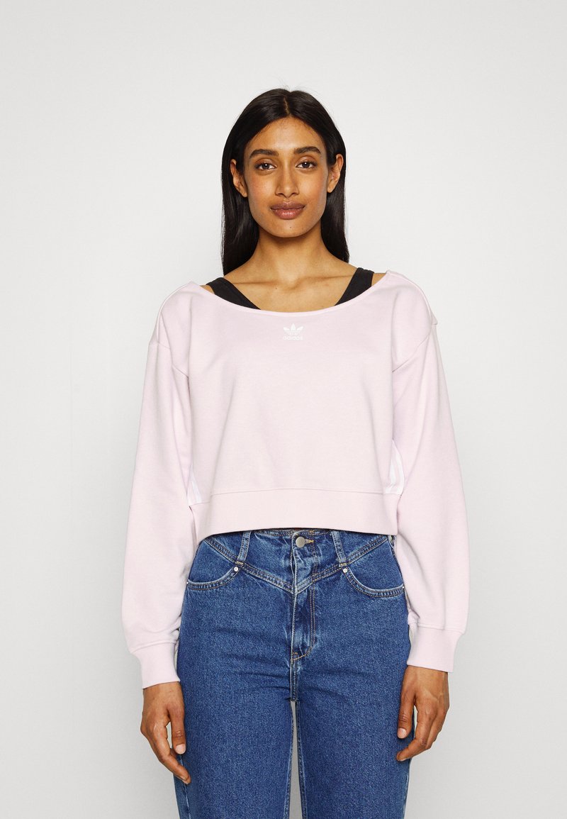 Sweatshirt cropped rosa claro com decote largo e mangas longas, apresentando um logótipo branco. Combinado com calças de ganga azuis de cintura alta.
