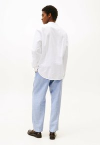 Camicia bianca a maniche lunghe con orlo arrotondato, abbinata a pantaloni a gamba larga a righe blu chiaro e mocassini scuri. Texture della stoffa liscia visibile.