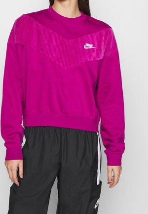 Personne portant un sweat-shirt Nike magenta vif avec un panneau chevron texturé et un pantalon Nike noir avec des bandes blanches sur les côtés.
