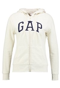 Sudadera cremosa con cremallera y letras en azul marino "GAP". Cuenta con capucha ajustable, dos bolsillos frontales y puños y dobladillo acanalados. Textura suave.
