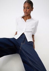 Chemise blanche à boutons avec les manches retroussées, assortie à un jean en denim bleu foncé à jambes larges avec des coutures orange contrastantes.
