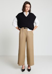 Schwarzer ärmelloser Pullover über einem weißen Hemd, kombiniert mit beigen Hosen mit weitem Bein. Schwarze High-Heel-Schuhe vervollständigen das Outfit.