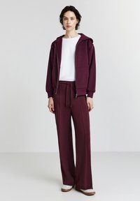 Felpa bordeaux con zip e tasche abbinata a pantaloni larghi coordinati, indossata sopra una t-shirt bianca a collo rotondo. Tessuto morbido e casual.