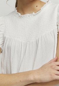 Vit topp gjord av texturerad, rynkad tyg med en volangkrage och korta ärmar. Plagget har en lös, avslappnad design.